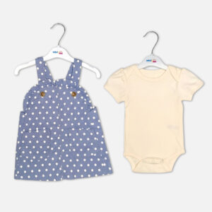 Baby girl 2-piece summer clothing set displaying a denim blue sleeveless pinafore dress with a white polka dot print, front patch pockets, and brown buttons, layered over a short-sleeve cream inner bodysuit from Mini & Me.