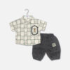Mini and Me boys two piece summer suit set hanging on a white hanger, featuring a cream short sleeve button down shirt with a green checkered grid pattern and a cartoon bear chest pocket graphic, paired with matching textured charcoal grey shorts featuring a small square number patch, displayed on a light gray background in file 20-1_2.jpg.