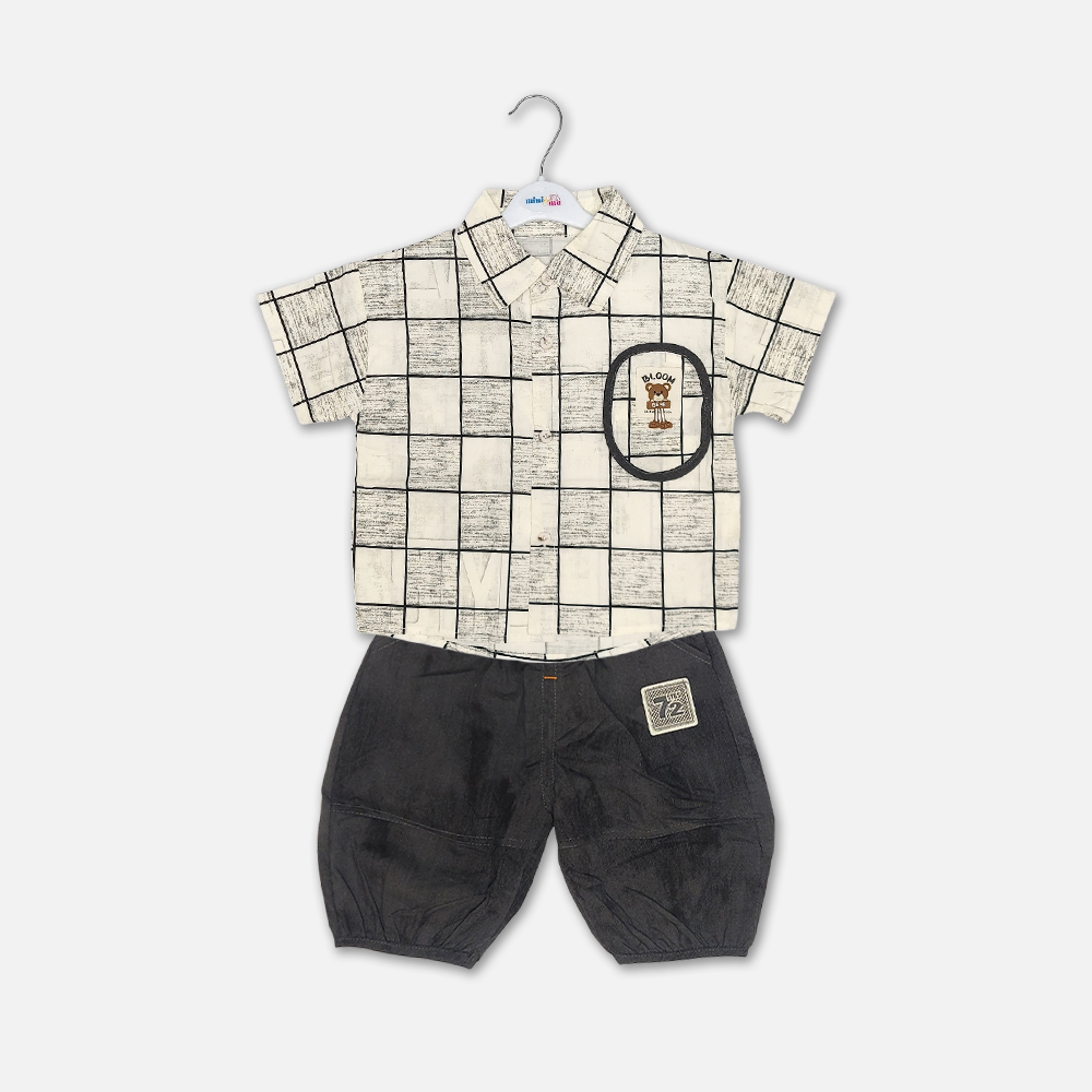 21-2 Mini and Me boys two piece summer suit set hanging on a white hanger, featuring a cream short sleeve button down shirt with a green checkered grid pattern and a cartoon bear chest pocket graphic, paired with matching textured charcoal grey shorts featuring a small square number patch, displayed on a light gray background in file 20-1.jpg.