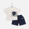 Mini and Me boys two piece summer suit set hanging on a white hanger, featuring a cream short sleeve button down shirt with a green checkered grid pattern and a cartoon bear chest pocket graphic, paired with matching textured charcoal grey shorts featuring a small square number patch, displayed on a light gray background in file 20-1.jpg.