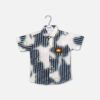 Mini and Me boys short sleeve button down shirt hanging on a white hanger, featuring a navy blue and white vertical striped pattern fused with large white tie dye cloud washes, an overlay of faint green letters, a structured collar, and a square brown faux leather chest patch, displayed on a light gray background.