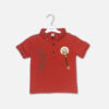 A premium rust red boys polo collar t-shirt with short sleeves, a three-button placket, embossed side lettering, and a round fashion patch with hanging accent cords, displayed on a hanger.