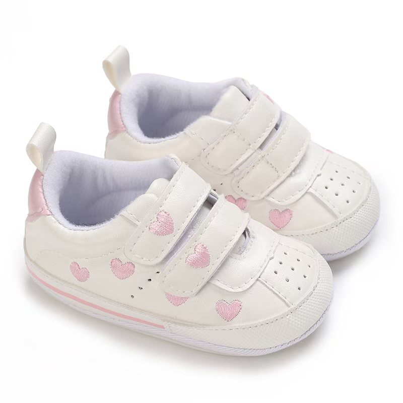 IMG-20260114-WA0115 Pair of white faux leather baby pre-walker sneakers featuring embroidered pink hearts on the sides and straps, dual adjustable hook-and-loop straps, a pink heel patch, and a flexible white outsole with a thin pink stripe.