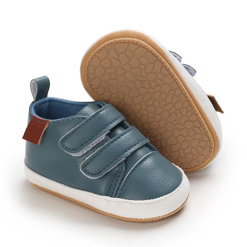 IMG-20260114-WA0120 Slate blue faux leather baby pre-walker shoes showing one shoe with dual hook-and-loop straps, a white midsole, and a brown side tag, alongside the other shoe flipped to display the tan, pebble-textured non-slip soft sole.