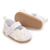 A pair of white infant dress shoes featuring an all-over floral lace overlay, a layered white fabric flower with a faux pearl center on the toe, a hook-and-loop instep strap, and a tan pebble-textured sole on a plain white background.