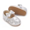 A pair of shimmering metallic silver infant dress shoes showcasing embroidered hearts on the toe box, a matching silver bow on the hook-and-loop strap, and a tan pebble-textured rubber sole on a plain white background.