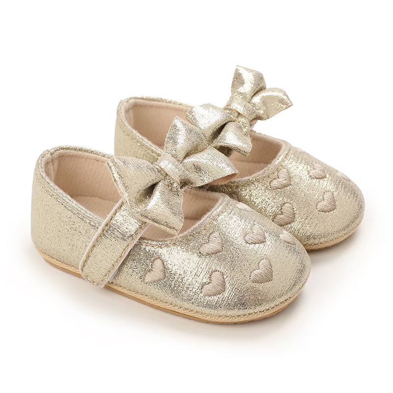 IMG-20260114-WA0135 A pair of shimmering metallic gold infant dress shoes showcasing embroidered hearts on the toe box, a matching gold bow on the hook-and-loop strap, and a tan pebble-textured rubber sole on a plain white background.