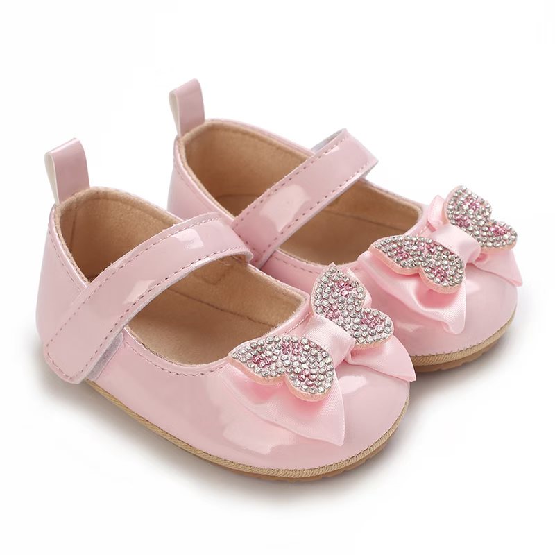 IMG-20260114-WA0137 Glossy pink baby girl shoes featuring a rhinestone butterfly bow embellishment and an adjustable hook and loop ankle strap, displayed against a clean white background.