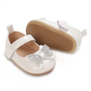 A pair of high-gloss white patent leather baby dress shoes showcasing a glittering rhinestone bow on the toe box, a secure hook-and-loop instep strap, and a tan pebble-textured rubber sole on a plain white background.