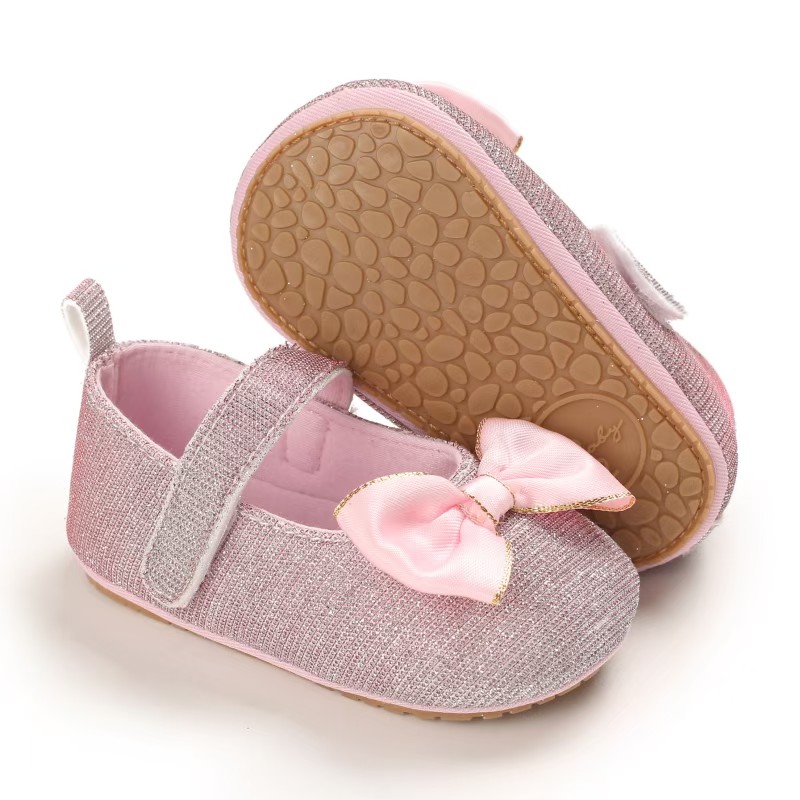 IMG-20260114-WA0144 Sparkly rose pink glitter baby girl shoes featuring a pink satin bow with gold trim, an adjustable hook and loop strap, and a textured tan non-slip rubber sole, shown on a white background.