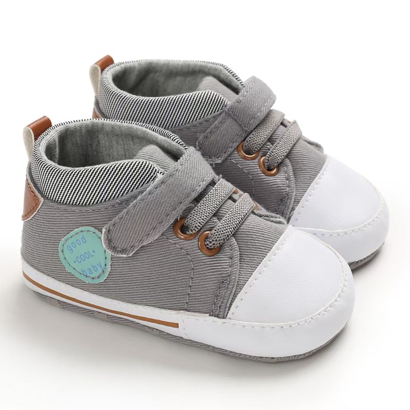 IMG-20260114-WA0154 Pair of grey canvas high-top baby sneakers featuring a white protective toe cap, grey elastic laces with brown metal eyelets, a single hook-and-loop strap, a round mint-green side patch, and a grey-and-white striped ankle collar.