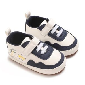 Pair of off-white leather baby sneakers featuring navy blue panels, a single hook-and-loop strap over white elastic laces, a navy blue inner lining, and a small cartoon dinosaur illustration with a speech bubble on the side.