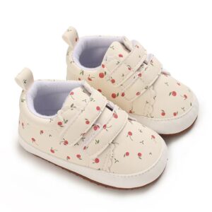 A pair of cream colored infant sneakers featuring an all-over red cherry and green leaf print, scalloped details on the double hook-and-loop straps, a white rubber-like toe cap, and a tan textured sole on a plain white background.