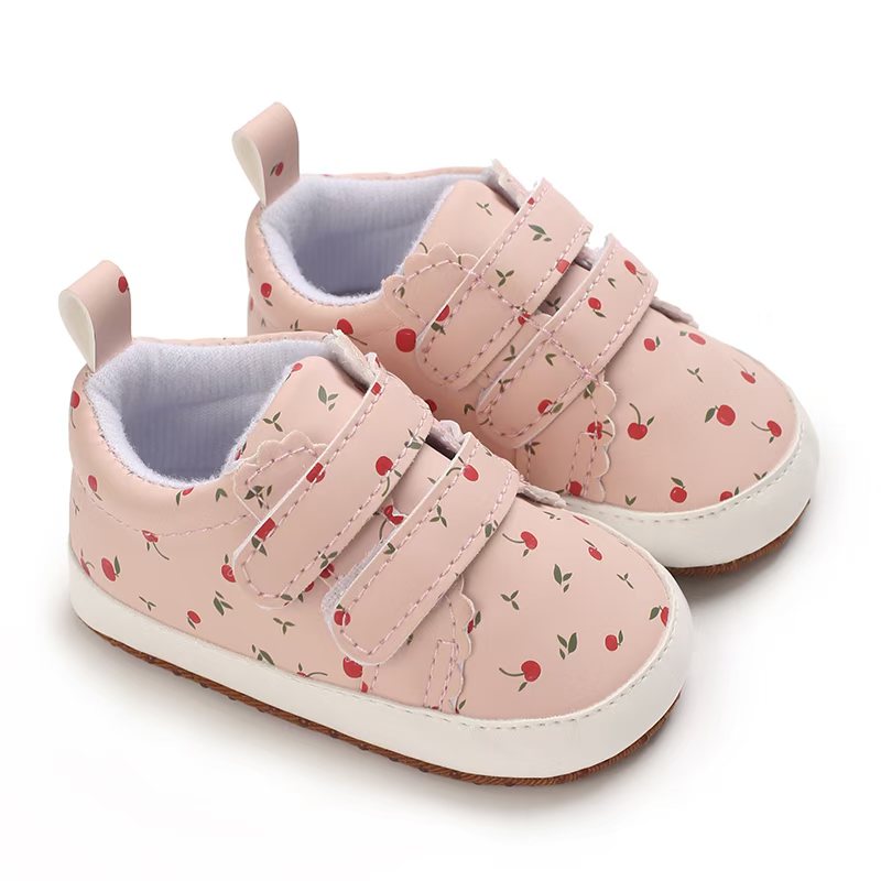 IMG-20260114-WA0163 A pair of pink colored infant sneakers featuring an all-over red cherry and green leaf print, scalloped details on the double hook-and-loop straps, a white rubber-like toe cap, and a tan textured sole on a plain white background.