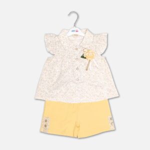 A premium two-piece summer clothing set for baby girls featuring a cream button-down blouse with an all-over ditsy floral print, ruffle sleeves, and a yellow fabric flower brooch, alongside pastel yellow shorts with matching floral side panels, displayed on a hanger.