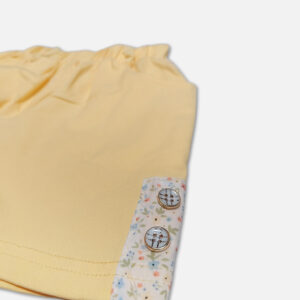 A premium two-piece summer clothing set for baby girls featuring a cream button-down blouse with an all-over ditsy floral print, ruffle sleeves, and a yellow fabric flower brooch, alongside pastel yellow shorts with matching floral side panels, displayed on a hanger.