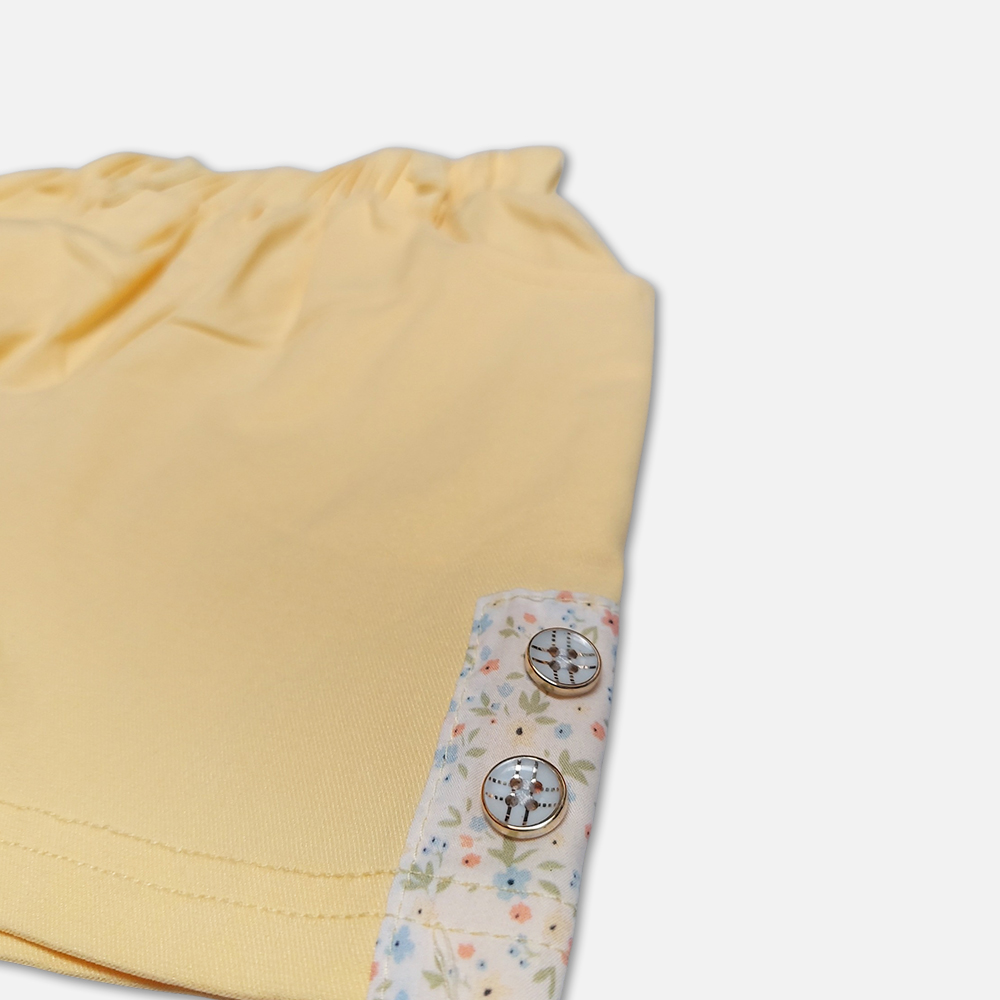 girlsSuit13134-2-6 A premium two-piece summer clothing set for baby girls featuring a cream button-down blouse with an all-over ditsy floral print, ruffle sleeves, and a yellow fabric flower brooch, alongside pastel yellow shorts with matching floral side panels, displayed on a hanger.