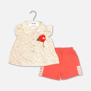 A premium two-piece summer clothing set for baby girls featuring a cream button-down blouse with an all-over ditsy floral print, ruffle sleeves, and a red fabric flower brooch, alongside coral pink shorts with matching floral side panels, displayed on a hanger.