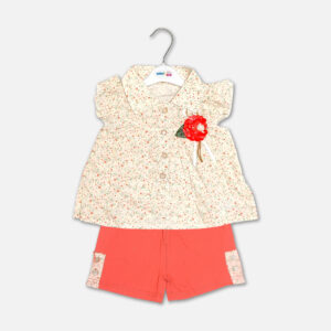 A premium two-piece summer clothing set for baby girls featuring a cream button-down blouse with an all-over ditsy floral print, ruffle sleeves, and a red fabric flower brooch, alongside coral pink shorts with matching floral side panels, displayed on a hanger.